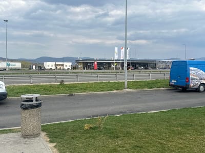 The image depicts an outdoor scene in Orăștie, Romania, at a commercial service area alongside a multi-lane highway, under a cloudy sky.
In the immediate foreground, a textured gray concrete trash receptacle with a black plastic bag liner visible at its opening is positioned on a paved surface, adjacent to a manicured grassy strip. A portion of a white vehicle is visible on the far left.
A two-lane paved road runs horizontally across the midground, separated from the commercial establishments by a silver metal guardrail. A bright blue van, featuring a white graphic print on its side and a visible Romanian license plate beginning with "HD", is parked facing away from the viewer on the right side of this foreground road.
Across the highway, a commercial complex is situated. This complex includes a dark-colored KFC restaurant with a drive-thru lane on the right. To its left, a larger building under a canopy structure is present, likely a gas station or convenience store, identified by multiple white flags bearing commercial logos (including "KFC" and what appears to be a fuel brand) mounted on poles. Numerous vehicles are parked or in transit within this area. Further left and deeper in the background, two white, modular-style rectangular buildings are visible. Several indistinct human figures are discernible near these buildings, indicating pedestrian activity. Beyond the commercial structures, a large white semi-truck with a green "for transit" logo on its trailer is visible on a parallel road or parking area. The distant background features rolling hills or mountains partially obscured by a cloudy sky.