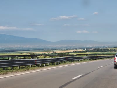 The image depicts a view from a highway, looking towards a distant city and mountains under a partly cloudy sky. The foreground shows a paved road with white lane markings, bordered by a metal guardrail. Beyond the guardrail, there are green grassy areas and fields, some appearing to be cultivated with crops. In the far distance, a city skyline is visible nestled at the base of hazy mountains. The city of Eleshnitsa, Bulgaria, is not identifiable in this image. A silver SUV is partially visible on the right side of the road. The overall scene is one of a rural or suburban landscape viewed from a major road.