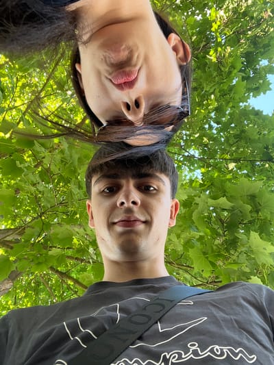 This is a low-angle, close-up, portrait-style selfie of two young people outdoors, with lush green foliage and a bright blue sky filling the background. The subjects are positioned at the top of the frame, looking upwards towards the camera.

On the left is a person with dark hair and what appears to be a portion of their face and lips visible. They are wearing dark-rimmed sunglasses perched on their head. Their lips are slightly parted, and their expression is somewhat serious or thoughtful.

To the right of this person is another individual, whose face is more fully visible. They have dark hair and are looking directly at the camera with a slight smile. Their right ear is visible. They are wearing a dark t-shirt with a white graphic design on it. A black strap with white lettering runs across their chest, obscuring part of the t-shirt's design.

The setting is clearly outdoors, under a canopy of vibrant green leaves, suggesting a park or wooded area. The bright lighting and clear sky indicate it is daytime, likely a sunny day. The angle of the shot and the way the subjects are positioned looking up suggest they are either lying down or standing very close to the camera, taking a selfie from a low vantage point. The overall mood seems relaxed and informal.

There is some visible text on the black strap worn by the person on the right. While the full text is not clearly legible, it appears to be a brand name or logo with repeating characters.