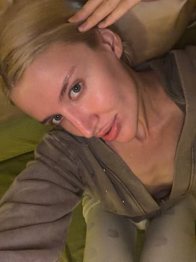 The close-up image features a fair-skinned individual, likely an adult female, with blonde hair and blue eyes, captured in a relaxed, intimate setting. The subject is lying down, positioned with their head on the left side of the frame (when viewed upright), and looks directly at the camera. One hand is gently placed near the ear, appearing to brush back hair. The lips are slightly parted, and the expression is calm and direct.
The person is dressed in a comfortable-looking, soft-textured hooded garment, possibly a jacket or sweatshirt, in a muted greyish-brown color. Small, metallic embellishments, resembling studs or beads, are visible on the shoulder or upper arm area of the garment. A lighter-colored piece of clothing or fabric is visible underneath or extending from the lower part of the body.
The scene appears to be indoors, suggesting a private environment like a bedroom or living room. A soft, olive-green fabric, possibly a blanket, pillow, or upholstery, is prominent in the background, contributing to the relaxed atmosphere. The lighting is soft and ambient, illuminating the subject's face, which shows a healthy, slightly glowing complexion. There is no visible text in the image. The overall impression is one of candidness and comfort in Nerin, Montenegro.