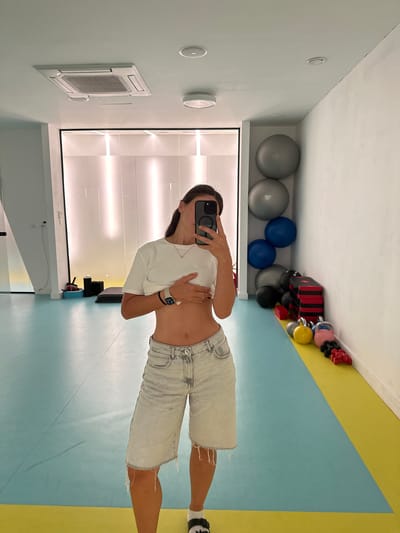 The image shows a woman taking a mirror selfie in what appears to be a gym or fitness studio. She is wearing a white t-shirt and light-colored denim shorts. The studio has light blue and yellow flooring. In the background, there are several exercise balls and some weightlifting equipment on a shelf. The walls are light blue and white. There is also a large air conditioning unit mounted on the wall. The lighting in the room is bright, suggesting it is daytime. The woman is holding her phone to take the picture, and her expression is neutral. There is no visible text in the image. The scene suggests a casual workout session. The location is Boreti, Montenegro.