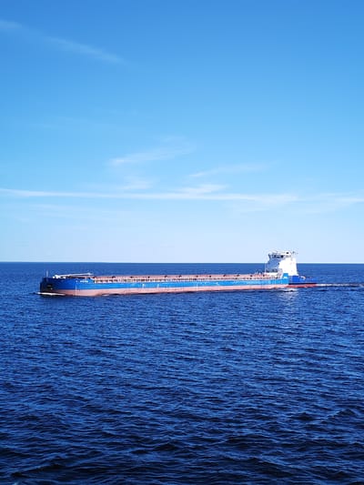 This image captures a cargo vessel, identified by the hull number "МУ 87368" and IMO number "7269046" (Rybinskaya-600), navigating a wide expanse of water under a clear blue sky, likely on the Rybinsk Reservoir near Мякса, Russia.
The vessel is a long, low-profile ship with a blue hull, a reddish-brown waterline, and a white superstructure located at its aft. It is positioned roughly in the center of the frame, moving from left to right, leaving a discernible white wake trailing behind its stern. The deck features white railings, and the cargo holds appear covered or empty.
The water is a deep cerulean blue, covered with numerous small, wind-induced ripples and waves, indicating light surface agitation. The horizon line is flat and clearly defined, separating the water from the sky. The sky above is a bright, uniform cerulean blue, with only faint, wispy white clouds scattered sparsely in the upper portion. The scene is devoid of other vessels, people, or visible landmasses in the immediate vicinity of the ship, portraying an open-water environment under sunny conditions.