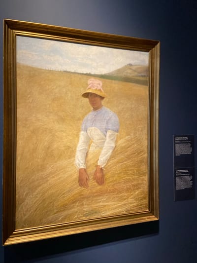 The image is a painting of a woman wearing a straw hat and a white dress with a blue jacket, resting in a field of wheat. The painting is framed in a gold frame and hangs on a blue wall. It is located in a museum in Greenwich, Connecticut, United States. The coordinates provided (latitude: 41.02, longitude: -73.623) are close to the Bruce Museum, which is known to have a collection of paintings.