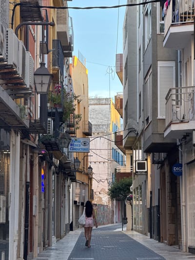 The image captures a narrow street in Vinaròs, Spain. Buildings line both sides of the street, with a mix of architectural styles and varying heights. A woman walks along the right side of the street, dressed in a light pink outfit, with a white bag or cloth over her shoulder. Overhead power lines are visible, crisscrossing above the street.

The buildings display an assortment of elements: air conditioning units, balconies, window blinds, and signs, including one that reads "CENTRO MEDICO CONDUCTORES" in Spanish. The street surface is composed of gray rectangular pavers. The overall atmosphere is urban and densely built.