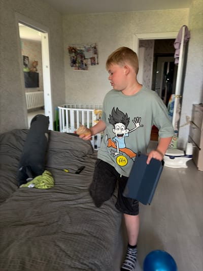 A young boy with blonde hair and a light green t-shirt with a cartoon character print is standing on a wooden floor in what appears to be a bedroom or play area. He is holding a dark blue tablet in his right hand and a light brown object, possibly an ice cream cone, in his left hand. He is wearing dark shorts and striped socks. In the background, there is a bed with a dark gray comforter, a long dark gray pillow, and a green cloth. A white crib is visible further back, along with a door leading to another room. A blue balloon is on the floor near the boy's feet. The room is decorated with wallpaper and framed pictures on the wall.
