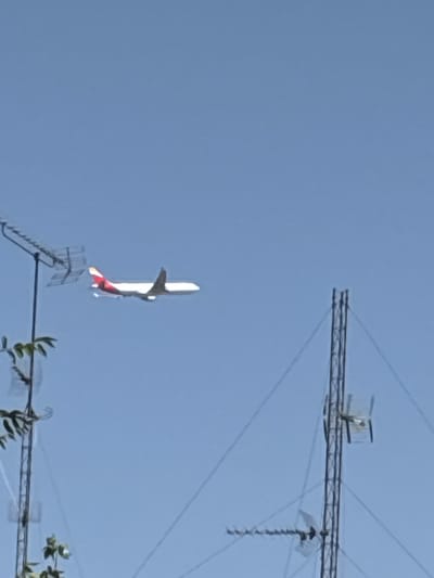 The image shows a model airplane with white wings and a red tail in flight against a clear blue sky. The airplane is positioned in the center of the frame, appearing to be in motion. In the foreground and upper right of the image, parts of what appear to be antennas or radio towers are visible. These structures are made of metal bars and wires, with some components resembling signal receivers. The sky is uniformly blue, suggesting a clear day, likely during daylight hours. There are no people visible in the image. The overall scene depicts an outdoor environment with the airplane as the primary subject, possibly being flown in an open area near telecommunication equipment. No text is visible in the image. The location context provided is Coslada, Spain.