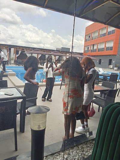 The scene takes place in a hotel courtyard in Ujevwu, Nigeria, with the coordinates provided. The view is from inside a building, looking out through a glass window. The courtyard features a swimming pool, patio furniture, and a large building with an orange facade. In the foreground, there are four people standing in the courtyard. Three women are positioned near the center, facing the camera. The woman in the center, wearing a floral dress, appears to be adjusting her hair. The woman to her left, wearing a white bikini top and black bottoms, is standing with her back to the camera, and the woman to her right, wearing a white dress, is looking towards the camera. The woman in the center is holding a black purse. To the left of the women stands another woman in a long white dress, black boots, and braids. She is looking down at a phone in her hand, which she is holding up to her face. A man in a white shirt and black pants is seen through the window in the background, standing on the far left side of the scene. He is wearing a white shirt and black pants. The ground is covered in white gravel and there is a large green couch in the foreground, obscuring part of the view. The blue swimming pool in the background is mostly obscured by the patio furniture. The building with the orange facade has multiple windows with white railings. There is an awning above the patio area, and there are palm trees in the background. There is a blue pool in the background. It is a sunny day.