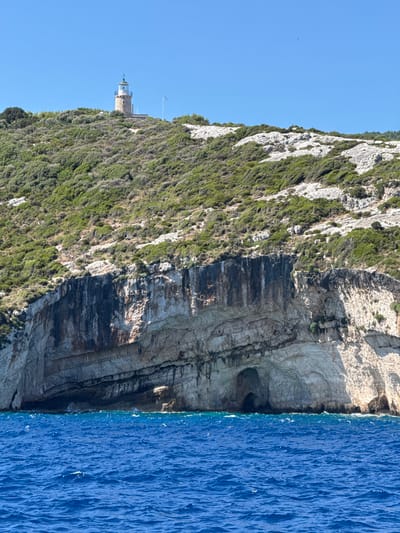 This image shows a view from the water towards a rocky, green hillside with a lighthouse on top. The hillside is covered in dense, low-lying vegetation and some bare rock. A large cave is visible at the base of the cliff face, which is made up of layered rock. The water in the foreground is a vibrant blue with small waves. The sky is clear and bright blue. The lighthouse, a stone structure with a white lantern room, is located on the highest point of the hill. A Greek flag is planted to the right of the lighthouse. This scene is consistent with the coastline of Elaties Municipal Unit, Greece, known for its dramatic cliffs and lighthouses.