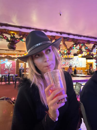 A blonde woman in a black cowboy hat drinks from a clear glass, her eyes looking upwards. She wears a black top with a fur collar. Christmas lights decorate a carved wooden structure in the background, next to a wall with framed pictures. In the lower portion of the frame are chairs and a painting on the wall.