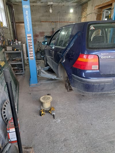 A blue Volkswagen Golf is lifted on a Twin Busch two-post lift in what appears to be a workshop or garage. The car is oriented with its rear facing the viewer, and the rear passenger-side wheel is missing, exposing the brake mechanism. Beside the car, a yellow and black impact wrench rests on a white spool. The environment seems to be an auto repair facility with various tools and equipment. The location could be Rozovo, Bulgaria, given the presence of the "herrnholz.de" sticker on the car which indicates a company that may be located in Germany or another German-speaking region.