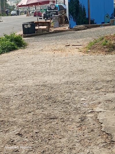 The image shows a dirt road with a few patches of grass on the side, taken from the perspective of a person standing on the road. The road is mostly brown and gray and appears to be in need of repair. In the background, there is a small shop with a blue awning. There are people walking on the road, and a car is parked in the distance. The scene is likely located on a street in Ado Ekiti, Nigeria, based on the provided coordinates. The only text in the image is a sign that says "Beverages, Drinks, Jewelries" and a watermark that says "Aramko, Nigeria" in the bottom left corner. The image was likely taken during the day, as the lighting is bright.