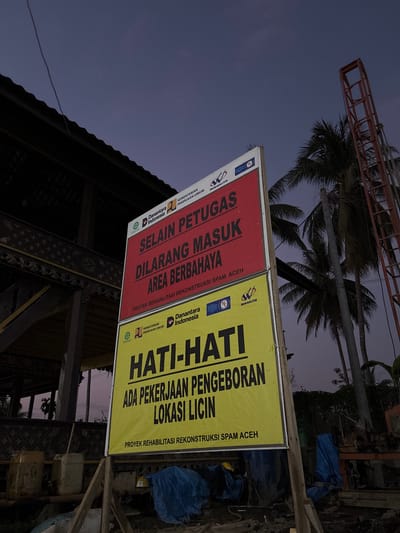 This media file captures an outdoor scene at what appears to be a construction or rehabilitation site in Mideuenjok, Indonesia, during dusk or early night, given the dark blue and purple hues of the sky. No people are visible.
The primary subjects are two large informational signs, mounted side-by-side on wooden posts. The left sign is predominantly red with white text, while the right sign is yellow with black text. Both signs bear logos for "Danantara Indonesia," "KEMENTERIAN PEKERJAAN UMUM" (Ministry of Public Works), and "WASKITA" (a construction company).
The red sign states: "SELAIN PETUGAS DILARANG MASUK AREA BERBAHAYA," which translates to "EXCEPT FOR OFFICERS, ENTRY TO DANGEROUS AREA IS PROHIBITED." Below this, it identifies the activity as "PROYEK REHABILITASI REKONSTRUKSI SPAM ACEH," meaning "REHABILITATION AND RECONSTRUCTION PROJECT OF SPAM ACEH" (likely referring to a Drinking Water Supply System).
The yellow sign reads: "HATI-HATI," meaning "BE CAREFUL" or "CAUTION." It further warns: "ADA PEKERJAAN PENGEBORAN LOKASI LICIN," translating to "THERE IS DRILLING WORK, SLIPPERY LOCATION." This sign also identifies the project as "PROYEK REHABILITASI REKONSTRUKSI SPAM ACEH."
In the background, several palm trees are silhouetted against the dim sky. Parts of a building structure, possibly a traditional wooden building with intricate carvings, are visible beneath the signs. On the ground near the yellow sign, blue tarps and some containers can be seen, hinting at ongoing construction or storage of materials. The overall scene conveys a cautionary message about an active construction or infrastructure rehabilitation project in a tropical setting.