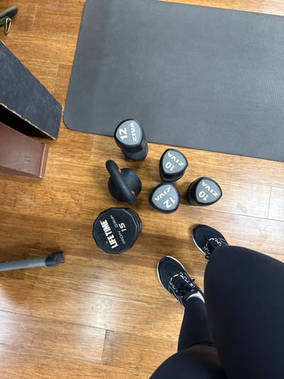 An overhead view depicts an indoor workout scene in Plainsboro Township, United States. The floor is composed of light brown wooden planks. A dark grey exercise mat is positioned in the upper right. Various free weights are arranged on the floor: one ZIVA 12-unit cylindrical dumbbell, two ZIVA 10-unit triangular dumbbells, two ZIVA 12-unit triangular dumbbells, one black kettlebell with red accents, and one black 'LIFETIME IRON GRIP 15' octagonal weight. The lower legs and feet of an individual are visible in the lower right, clad in black athletic leggings and black athletic shoes with white soles and lacing. In the upper left, a dark-colored wooden or composite object is partially visible, and a grey metal pole lies on the floor in the lower left.