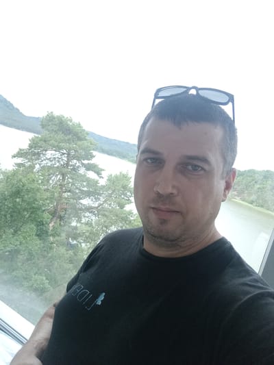 A man, likely in his late 30s or early 40s, is taking a selfie in front of a window. He has short, dark hair and is wearing a black t-shirt with a light blue logo on the left side of his chest. He has sunglasses resting on the back of his head. The background reveals a scenic view of a body of water, possibly the Mácha Lake, surrounded by lush green trees and hills. The sky is overcast, suggesting it might be a cloudy day. The man is positioned in the foreground, with his face and upper body filling most of the frame. The window he is standing in front of appears to be large, offering an unobstructed view of the outdoors. The location appears to be in or near the town of Doksy, Czechia, known for its natural beauty and the popular Mácha Lake.