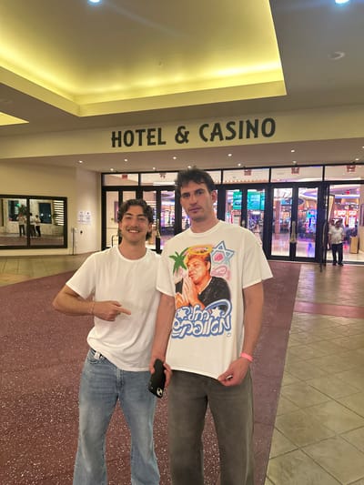 Two young adult males are posing in front of the entrance to a casino. Above them is the signage "HOTEL & CASINO". The casino entrance is visible in the background. The location is the Seminole Hard Rock Hotel & Casino in Hollywood, Florida, USA. The male closest to the camera is wearing a plain white t-shirt and blue jeans, pointing towards the other male, who is wearing a white graphic t-shirt and grey pants, and holding a mobile phone.