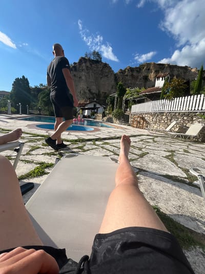 In Melnik, Bulgaria, a sunny day unfolds with a view of a pool area. A bald man in a black t-shirt and shorts walks away from the camera towards a circular pool where pink inflatable objects are visible. The pool is surrounded by a stone patio with grass growing between the stones. In the foreground, the legs of a person lying on a lounge chair are visible. Behind the pool, there's a white picket fence, a stone building with a tiled roof, and a rocky cliff face. The sky is blue with scattered white clouds.