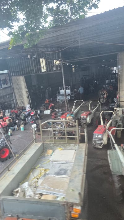 The image depicts a cluttered scene inside what appears to be a vehicle repair shop or storage facility in Pơng Drang, Vietnam. The location is identifiable by the license plates on the motorbikes. Various mechanical equipment, including several small tillers or cultivators, are visible. Some of the machinery is located inside the building, while other units are being loaded onto the back of a truck. The floor and surrounding areas are dirty and appear to be wet, suggesting a recent cleaning or rain. The building has a corrugated metal roof and siding, and there is foliage from nearby trees visible on the left side of the frame.