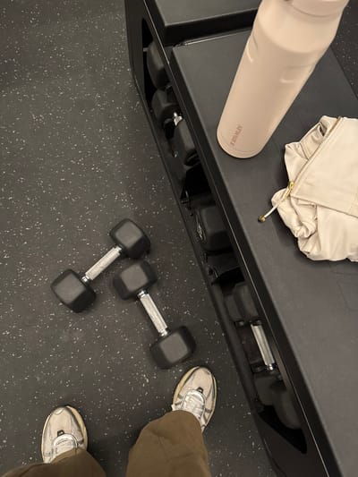 The scene depicts an interior view of a fitness facility in Dudley, United Kingdom, captured from an overhead, first-person perspective looking downwards. The floor is dark grey, speckled rubberized gym matting. In the foreground, the lower legs and feet of an individual are visible, wearing olive-brown trousers and light grey athletic shoes with white laces and accents. Two black hexagonal dumbbells with silver handles rest on the floor directly in front of the individual. To the right, a dark, multi-tiered dumbbell storage rack is present, partially filled with additional dumbbells on its lower shelves. The top surface of this rack holds a light pink Stanley brand water bottle lying horizontally and a folded, light-colored (off-white/cream) athletic jacket featuring a gold zipper pull and a matching drawstring. No individuals are depicted beyond the visible lower extremities.