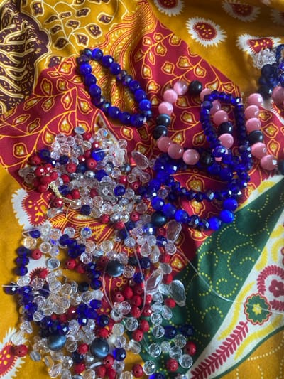 The image presents a top-down view of various beads and partially assembled beadwork displayed on a multi-colored patterned fabric. The fabric features distinct sections with designs in yellow, red, maroon, brown, and green, suggesting a traditional textile. In the upper portion, several strands of beads are visible: one strand of large, faceted royal blue beads, multiple strands of alternating opaque pink and dark blue/black round beads, and multiple strands of smaller faceted royal blue beads. The lower and central left areas are occupied by a dense cluster of loose, unstrung beads. This pile primarily consists of small, round, opaque red beads; clear faceted beads of various sizes and shapes (including round and teardrop); and smaller faceted royal blue beads. Interspersed within the loose beads are several larger, opaque, dark blue/black round beads, some exhibiting a cat's eye effect, and strands of transparent monofilament beading thread, indicating an ongoing or preparatory crafting process. The scene is located in Odo Ona, Nigeria.