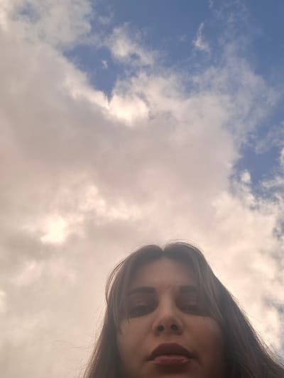 A selfie of a person with long, brown hair with visible red lipstick is in the lower-left corner of the frame. The person's head is tilted to the side. The background is a partly cloudy sky with sections of blue. The location cannot be confirmed.