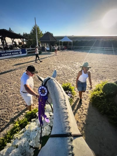 The image captures an outdoor equestrian arena at Thunderbird Show Park in Langley, Electoral Area A, British Columbia, Canada, under a clear, bright sky. The perspective is taken from just behind and above a light-colored horse, looking forward over its head and braided neck. The horse's bridle is adorned with a large purple and white championship rosette and ribbons.
To the left of the horse, a man wearing a white t-shirt, white shorts, black baseball cap, and sunglasses stands, his right hand positioned near the horse's head. To the right, a woman wearing a white tank top, light-blue denim shorts, and a wide-brimmed light-colored hat walks forward on the sandy arena surface, looking towards the horse.
The immediate foreground and a path extending into the midground are bordered by decorative elements consisting of dense white floral arrangements and green foliage, including some large spherical topiary-like bushes. The arena floor is composed of light-colored sand or dirt.
In the background, to the left, is a large, dark-roofed, open-sided structure with a banner attached below it that reads "WORLD CHAMPIONSHIP HUNTER RIDER" and "UNITED STATES HUNTER JUMPER ASSOCIATION." To the right of this structure, a smaller dark-roofed kiosk and a white peaked tent are visible within the arena perimeter. A white fence or railing defines the far edge of the arena, beyond which a darker treed or vegetated landscape is present. Long shadows are cast across the arena surface from the left, indicating the sun's low position.