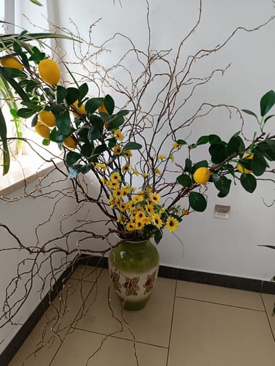 This is a medium shot, eye-level, indoor photograph of a decorative floral arrangement in a green vase. The arrangement features artificial lemons on branches with green leaves, intertwined with bare, brown branches. Small yellow flowers are also incorporated into the bouquet. The vase is positioned on a tiled floor against a white wall. The setting is likely a domestic or commercial interior, but there is no specific information to pinpoint it to Czekanów, Poland. There are no people or animals present in the image.