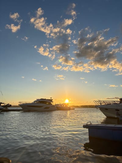 The image depicts a harbor scene at sunset in Tønsberg, Norway. The sky is a gradient of blue and orange, with scattered white and gray clouds illuminated by the setting sun. The sun, a bright orb, is positioned on the horizon, casting a warm glow across the water and the sky. Several yachts are moored in the harbor. In the foreground, the rippling surface of the water reflects the colors of the sunset. On the right side of the frame, a portion of a white yacht is visible, its metallic railings catching the light. To the left, a larger white yacht is docked, its sleek design prominent. Further left, a sailboat mast is partially visible. The background shows a distant tree line, silhouetted against the sunset. The scene is calm and serene, capturing the beauty of a coastal evening.