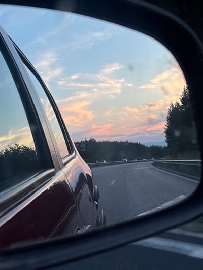 The image captures a sunset viewed through the side mirror of a car, likely on a highway near Baliovtsi, Bulgaria. The mirror reflects a dusky sky with streaks of orange, pink, and blue clouds, set against the dark silhouettes of distant trees and mountains. In the foreground, the metallic red body of the car is visible, curved and reflective. The road ahead is paved, with lane markings and a guardrail visible. Several cars are spaced out on the road, appearing as distant white specks. The overall impression is one of travel during a picturesque twilight.