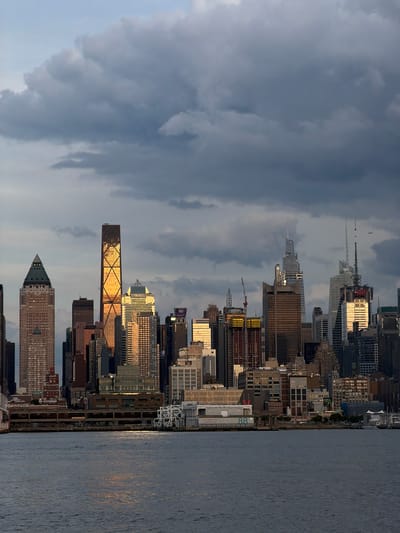 A panoramic view of the New York City skyline is captured from Weehawken, New Jersey, USA, across the Hudson River. The scene is dominated by a dense collection of skyscrapers, with the golden hue of the setting sun illuminating some of the buildings. Dark, dramatic clouds fill the upper portion of the frame, creating a contrast with the bright city lights. The foreground features the dark, rippling surface of the Hudson River, reflecting the sky and the distant structures. Along the waterfront, several buildings and structures are visible, including what appear to be piers and docks. A faint airplane can be seen in the sky above the skyline. The overall atmosphere is one of urban grandeur and the dramatic interplay of natural light and man-made structures.