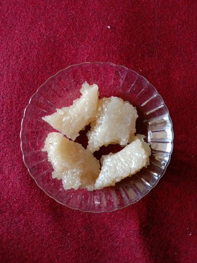 A top-down, medium shot depicts a clear glass plate containing four pieces of an unidentified, light-colored food item. The food pieces appear to be somewhat translucent and crystalline in texture. The plate has a fluted or ribbed edge. The plate rests on a dark red fabric surface. The image was taken in Blangkarieng, Indonesia.