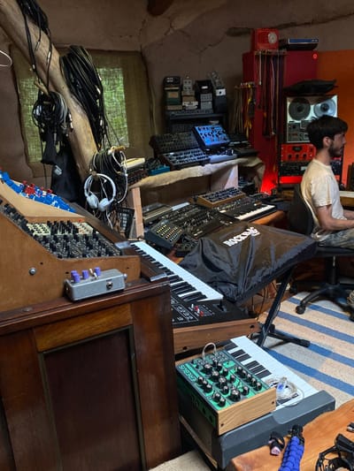 A cluttered room packed with synthesizers, modules, and audio equipment. A person sits in a chair facing a wall filled with gear. Cables, headphones, and various devices are scattered around the room. The room appears to be a studio or workspace dedicated to music production and electronic sound design. A "Rockbag" cover partially obscures a keyboard.