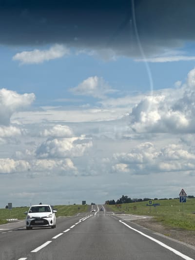 The image depicts a road in a rural setting. The perspective is from a vehicle traveling on the road. The road is asphalt with white markings and appears to be a two-lane highway. To the right of the road, there is a grassy shoulder and beyond that, fields of greenery and some trees. Several road signs are visible along the right side of the road, including directional signs and a sign with a cross, indicating a church or religious site. In the distance, there are more cars on the road.
In the foreground, a white car is visible, driving on the road. The license plate is partially obscured but readable as "5321KA-3". The sky is a mix of blue with scattered white clouds and a darker, possibly stormy cloud formation on the left side of the image, with a faint rainbow arc visible. This suggests a varied weather condition. The lighting indicates it is daytime.
The scene appears to be in a rural area, likely Belarus given the context provided. The overall impression is one of a journey through a landscape with an approaching change in weather.