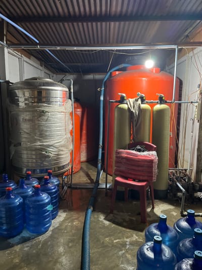 In the city of Teupinkuyun, Indonesia, a dimly lit storage room contains equipment for water purification and distribution. A large, stainless steel water tank labeled "T.1000" is covered in plastic wrap, situated next to two tall, orange water tanks. In front of these tanks are three vertical, olive-green filter tanks connected by pipes and valves. A red plastic stool, wrapped in plastic, holds a bundle of red tubing. The floor, made of concrete, appears wet and reflects the overhead lighting. Several blue water jugs with red caps are arranged in the foreground, and a blue corrugated pipe snakes across the floor. The ceiling is constructed of corrugated metal sheeting.