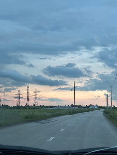 The image captures a roadside view in Qarabalıq, Kazakhstan, likely during twilight due to the soft, diffused light and the colors in the sky. The scene is dominated by a vast, cloudy sky with hues of blue, gray, and hints of pink and yellow, suggesting either sunrise or sunset.
A paved road, marked with white lines, stretches into the distance, curving slightly to the right. The road appears to be in moderate condition with some patches and a visible pothole. To the left of the road, there is a grassy embankment leading to a field. Several power lines and transmission towers are visible, stretching across the landscape and indicating the presence of infrastructure. One prominent transmission tower, made of lattice metal, stands tall in the middle ground.
In the background, a few scattered buildings, including what appears to be a white house with a dark roof, are visible amidst trees and vegetation. The overall atmosphere is serene and slightly melancholic, characteristic of a quiet evening or early morning in a rural or semi-rural setting. No people are visible in the image, and there is no discernible text. The orientation of the image is landscape, but it appears to have been captured sideways and may need to be rotated for proper viewing.