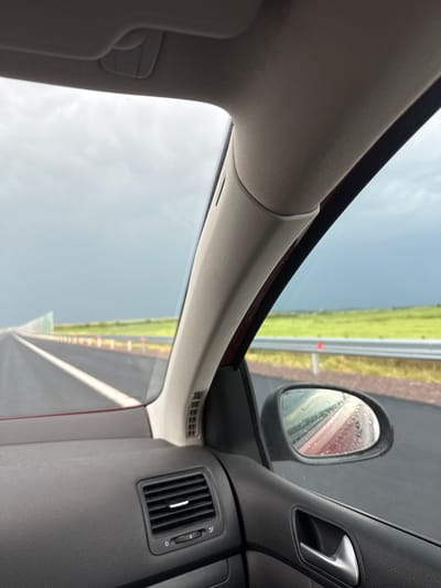 An eye-level interior shot from a vehicle captures a dynamic view, rotated ninety degrees clockwise, from the passenger side in Chokoba, Bulgaria. The car's A-pillar and part of the headliner, upholstered in light beige fabric, frame the left side and top of the image. A light-colored sun visor hangs partially into view on the far left.

Through the car's windshield and passenger window, an overcast sky is visible, with faint water droplets adhering to the glass, suggesting recent or ongoing rain. Beyond the window, a green, grassy expanse runs alongside a grey road or highway, which appears vertically oriented due to the image's rotation. Parts of a barrier or fence are also discernible along the road.

The right side of the image reveals elements of the car's dark grey dashboard and door panel. A textured black air vent with a vertical slider, faintly marked "0" and "1," is visible on the dashboard. Above it, a metallic door handle is integrated into the door panel. The side mirror, covered in numerous water droplets, shows a blurred reflection of the wet environment and a reddish hue, likely from the car's body. The overall scene depicts a journey in wet conditions, viewed from inside the moving or parked vehicle. No people are visible.