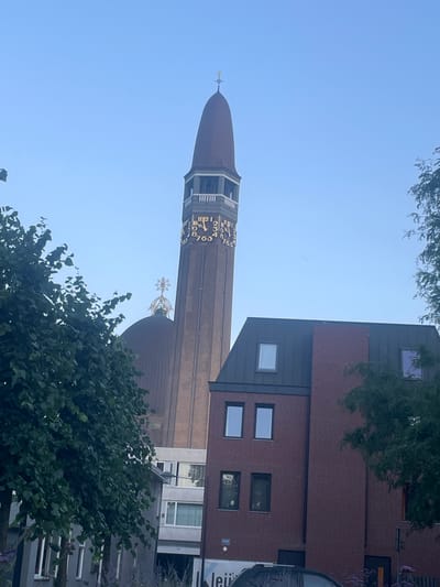 The image shows the St. Joseph's Church tower in Waalwijk, Netherlands, a tall brick structure with a conical copper-colored spire. A clock face with Roman numerals is visible on the tower. In front of the church, there is a modern brick building with several windows. Green trees partially obscure the view of the church and the building. The sky is clear and blue.
