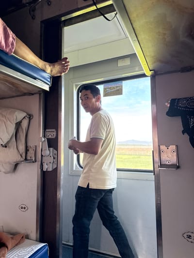 The image shows the interior of a train compartment with a man in a light-colored polo shirt and dark jeans walking towards the camera. He is looking back over his shoulder. To the left of the man, a person's bare foot is visible resting on a blue bunk bed. The compartment has multiple bunks, with blankets and bags stored on them. A window to the right reveals a bright, sunny outdoor scene with a green field and distant mountains. The train appears to be in motion, as suggested by the landscape outside. The overall impression is of a functional and somewhat utilitarian train travel experience. The location is identified as Bình Nguyên, Vietnam.