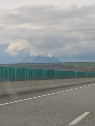 The image shows a highway in Konstantinovskaya, Russia. A mountain partially obscured by clouds is in the background. A green sound barrier runs along the side of the highway. A white line divides the lanes of the gray asphalt road. The sky is overcast.