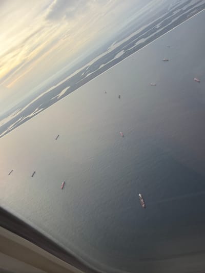 The image, taken from an aircraft window, shows a body of water filled with numerous ships. The coastline, marked by several narrow strips of land, is visible on the left side of the image. The water appears calm, and the ships are spread out across the frame. The lighting suggests it may be either sunrise or sunset. The angle of the shot and the presence of the aircraft window indicate the scene is viewed from an aerial perspective. The location is Communauté rurale de Diass, Senegal.