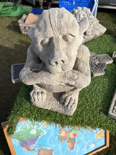 A high-angle, full-color photograph shows a collection of decorative items, potentially for sale, resting on the ground in Chirk, United Kingdom. A light gray, concrete-like gargoyle statue with folded arms is prominently positioned in the foreground, adjacent to a patch of artificial turf. To the right of the statue and turf is a framed world map with colorful continents on a blue ocean background. Other concrete decorations of varying shapes and sizes are scattered around, along with a blue plastic crate. The ground appears to be a grassy or dirt surface, suggesting an outdoor setting. The scene is captured in daylight, with the sun casting shadows on the objects.