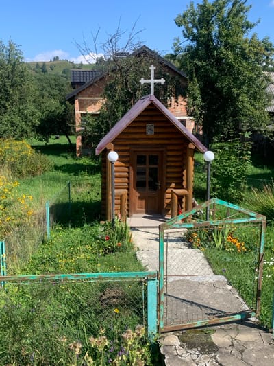 This image depicts an outdoor scene in Lazeshchyna, Ukraine, on a clear, sunny day. The central subject is a small, single-story structure constructed of horizontal dark brown logs, identified as a religious shrine or chapel. It features a sloped, dark-colored roof topped with a white Latin cross at its peak. A brown wooden door with a multi-paned window is centered on the front facade, and a small framed icon is positioned directly above the door. Two spherical white lamp globes on dark poles flank the entrance. To the immediate right of the door, a small, rustic wooden stand is present.

A concrete pathway, showing some cracking, leads from the foreground towards the structure's entrance. This path is bordered by a low, green-painted metal fence with chain-link mesh, which exhibits rust and chipped paint. An open gate, matching the fence's material and color, is positioned at the start of the path, swinging towards the viewer's right.

The surrounding environment is characterized by abundant vegetation: lush green grass, interspersed with various wildflowers in shades of yellow, orange, red, and purple, particularly dense to the left of the path. Numerous trees, some with full green foliage and others with visible bare branches, surround the structure and fill the midground. In the background, a two-story brick building with a dark roof is partially visible through the trees. Beyond this, rolling green hills or mountains rise under a clear blue sky with minimal cloud cover. No individuals or animals are present within the frame.