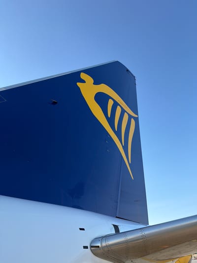 This media file is a close-up, low-angle shot of the tail fin of an airplane, likely at an airport given the bright sunlight and clear blue sky suggesting fair weather. The dominant colors are the deep blue of the airplane's tail fin, adorned with a stylized yellow bird or wing logo, and the bright, reflective silver of another part of the aircraft, possibly an engine nacelle or another section of the fuselage. The tail fin is angled, with the yellow logo clearly visible against the blue. The logo appears to be a stylized representation of a bird in flight, with multiple wing-like elements. The aircraft's surface shows a pattern of rivets and panel lines, indicative of its construction. On the blue tail fin, there are some small, dark rectangular shapes and circular indentations, likely access panels or sensors. The overall impression is one of a modern aircraft, possibly on the ground, awaiting departure or having just arrived. The lighting suggests it is daytime, likely midday, given the harsh shadows and bright illumination. There are no people visible in the frame. The location context provided is Luqa, Malta, which is known for its international airport, aligning with the subject matter. No text is visible in the image.