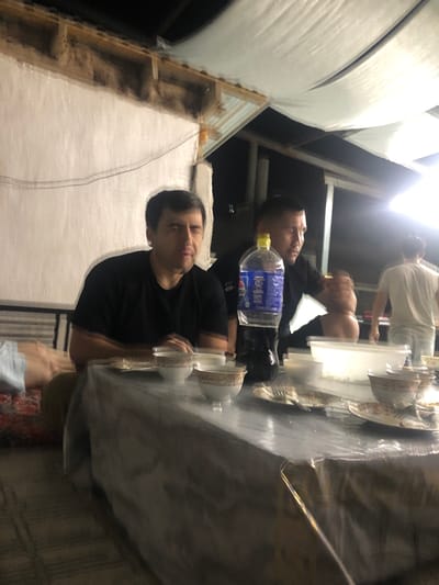 Two men are seated at a table set for a meal. The man on the left, wearing a black t-shirt, is looking forward with a neutral expression. The man on the right, also in a black t-shirt, is interacting with something on the table, his head turned down. A large bottle of Pepsi is on the table, along with several bowls and plates with food remnants and cutlery. In the background, another man in a light-colored t-shirt walks away from the table. The scene appears to be outdoors, under a makeshift roof structure covered with white fabric. The environment suggests a casual outdoor dining setting, possibly in Khujakent, Uzbekistan, given the context provided. The lighting is dim, indicating it may be evening or night.