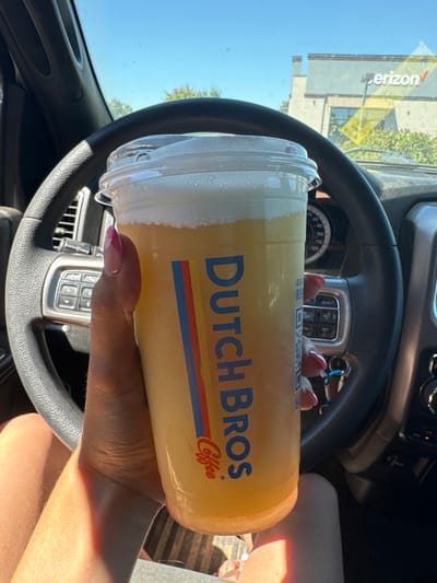 Inside a stationary vehicle in Granite Bay, California, United States, a person's left hand holds a large clear plastic Dutch Bros Coffee cup. The beverage is yellow-orange with a white foam topping. The hand exhibits a French manicure with pink and white polish. Visible vehicle components include a black steering wheel with integrated controls, a dashboard, and keys in the ignition. Through the windshield, a commercial building with 'Verizon' signage is visible, consistent with a parking lot at 5334 Sunrise Boulevard. The sky is clear and blue, with some distant trees also present.
