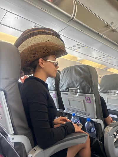 The scene depicts the interior of an airplane cabin, situated in the city of Las Veredas, Mexico. A female individual is seated in a gray airline seat, viewed in profile from the mid-torso up, facing towards the right side of the frame. She wears a dark, long-sleeved collared shirt and white-framed sunglasses. Her hair is pulled back, revealing an ear with a small stud earring. Perched prominently on her head is a stack of three straw hats of varying shades of brown and tan, the uppermost hat featuring a decorative geometric band. In front of her, the back of another airline seat is visible, featuring a gray headrest cover, a seat-back tray table in the folded-up position, and a small display area with a purple QR code and "Volaris +" branding. Two clear plastic water bottles, labeled "pura," rest upright on the folded tray table. A portion of another passenger's arm is faintly visible in the seat directly in front of the primary subject. The overhead panels of the aircraft cabin are visible in the upper portion of the frame, showing reading lights, air vents, and general cabin lighting fixtures.