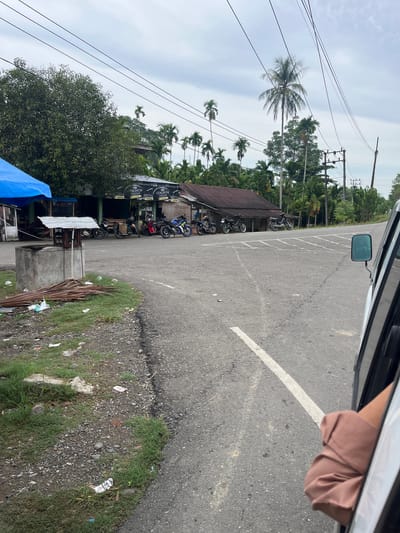 A low-angle shot captures a roadside scene in Jok Tanjong, Indonesia. The image is oriented vertically, with the sky taking up the top half and the road occupying the bottom. A person's arm, clad in a light brown sleeve, is visible resting on the car door, suggesting the viewpoint is from inside a vehicle.

The scene depicts a street lined with palm trees and dense foliage. Several motorcycles are parked in a row next to a long, low building with a dark roof. The building appears to be a small shop or eatery, with signs that read "MAGNUM FILTER" and prices like "25,000". A blue tarp is visible on the left side of the frame, possibly covering goods or a stall. The road itself is asphalt, marked with white lines. The ground beside the road is covered in gravel, dirt, and patches of green grass, with some litter scattered around. Utility poles with numerous wires are present in the background, indicating an inhabited area. The sky is overcast, suggesting a cloudy day. The overall impression is a typical rural or semi-urban street in Indonesia. No specific activity is clearly depicted, but the presence of parked motorcycles and the building suggests it might be a place of business or a resting stop.