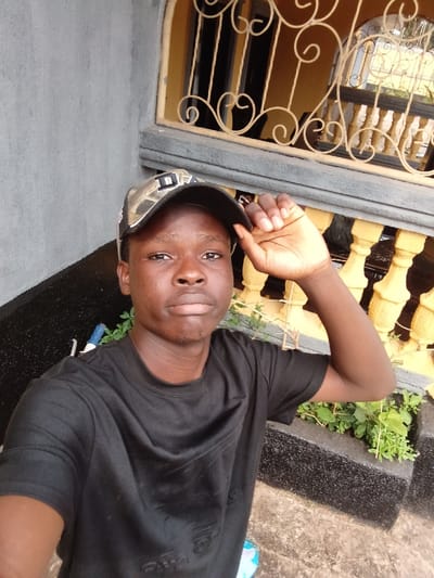 This image captures a young male individual standing outdoors in the city of Itu, Nigeria. The subject, with dark skin and short dark hair, is wearing a black short-sleeved t-shirt and a camouflage-patterned baseball cap with a dark brim, which displays a partial white letter or graphic on its front. His left hand is raised to the brim of his cap, and he looks directly forward with a neutral gaze.
The immediate environment includes a grey concrete or plastered wall to the subject's left. To his right and behind him, a building structure features a window or balcony with light yellow, possibly painted, balusters and an ornate cream-colored metal grille with scrollwork. A dark grey concrete ledge is situated below this architectural feature. Below the ledge, dark-colored raised planters contain green foliage and weeds. The ground surface in the foreground is concrete.
