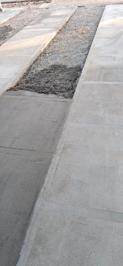 The image depicts an outdoor scene, possibly a construction site or a pathway under development, in the city of Mary, Turkmenistan. The foreground is dominated by a textured concrete surface, segmented into rectangular sections by lighter gray lines. This concrete appears to be laid out in a path or walkway. To the left of the concrete path, there is a trench or channel filled with gravel and some darker, possibly wet, material, suggesting recent work or preparation for something. This trench is bordered by concrete. In the background, more gravel and concrete structures are visible, along with what appears to be a wooden plank. The lighting suggests it is daytime, with shadows indicating sunlight. No people are visible in the image.
