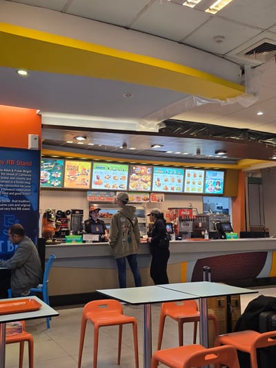 The image shows the interior of a fast-food restaurant, possibly a A&W franchise, located in Ramunia Dua, Indonesia. The restaurant features a brightly lit counter area with digital menu boards displaying food items and prices. Several staff members are visible behind the counter, serving customers. Customers are seen ordering and waiting for their food. The seating area includes orange plastic chairs and tables. The overall atmosphere appears to be a typical fast-food dining environment. The location is suggested by text on a sign that reads "Ramunia Dua, Indonesia."
