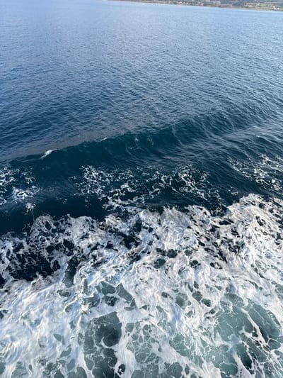 The image displays a maritime scene from an elevated perspective, featuring the wake of a vessel against a distant coastline. The lower portion of the frame is dominated by intensely churned, deep blue water, heavily aerated into white, frothy foam. This turbulent area transitions into undulating dark blue water with distinct waves in the mid-ground. The upper two-thirds of the image consists of calmer, lighter blue water with visible surface ripples. In the far background, a low-lying landmass is present, identified as the coastline of Kamariotissa, Greece. This distant land exhibits faint, indistinct low-rise structures and sparse vegetation. No individuals or identifiable specific objects are present within the immediate water scene. The scene suggests ongoing maritime travel in the vicinity of Kamariotissa.